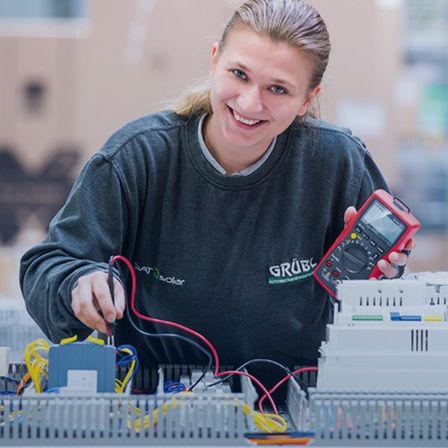 Eine Frau arbeitet in einer Fabrik, lächelt und hält ein Multimeter in der Hand, mit Kabeln und elektrischen Komponenten um sie herum.