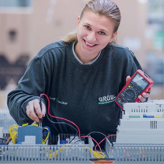 Eine Frau arbeitet in einer Fabrik, lächelt und hält ein Multimeter in der Hand, mit Kabeln und elektrischen Komponenten um sie herum.