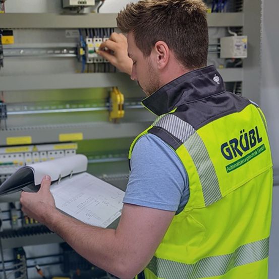 Ein Mann in einer gelben Sicherheitsweste mit der Aufschrift 'GRUBl' arbeitet an elektrischen Geräten und hält eine Ordner und Papier in der Hand.
