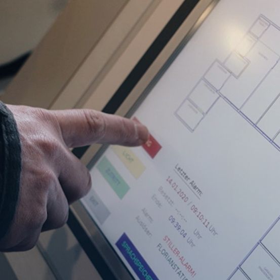 Eine Hand drückt einen roten Knopf auf einem Touchscreen, der eine Sicherheitsalarm-Benachrichtigung vom 14. Januar 2020 anzeigt. Der Alarm lautet 'Briefalarm' mit einem Zeitstempel von 09:39:04. Der Touchscreen zeigt auch den Namen 'Florian' und einen Status 'Sprechspreicher'.