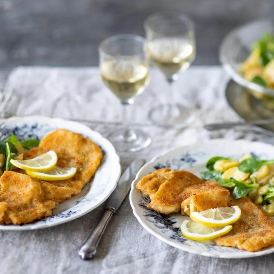 Zwei Teller mit frittierten Speisen mit Zitronenscheiben und Weingläsern auf einem Tisch. Ein Teller hat ein Messer daneben.