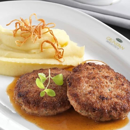 Zwei Fleischpatties mit grüner Garnierung auf einem Teller, begleitet von Kartoffelpüree und brauner Sauce.