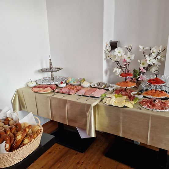 Ein Tisch mit goldener Tischdecke zeigt eine Vielzahl von Lebensmitteln, darunter Fleisch, Käse und Brot. Es gibt Blumen in einer Vase auf dem Tisch, und links steht ein Korb mit Brot.