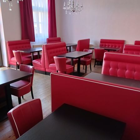 Ein Restaurant mit roten Leder-Booths, schwarzen Tischen und Kronleuchtern. Die Wände sind weiß und es gibt ein Fenster mit roten Vorhängen.