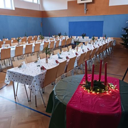 Ein Saal mit langen Tischen und Stühlen, die für einen festlichen Anlass gedeckt sind. Dekorationen umfassen rote Kerzen, einen Kranz und Weihnachtslichter. Der Raum hat blaue Wände und Holzböden.