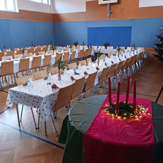 Ein Saal mit langen Tischen und Stühlen, die für einen festlichen Anlass gedeckt sind. Dekorationen umfassen rote Kerzen, einen Kranz und Weihnachtslichter. Der Raum hat blaue Wände und Holzböden.