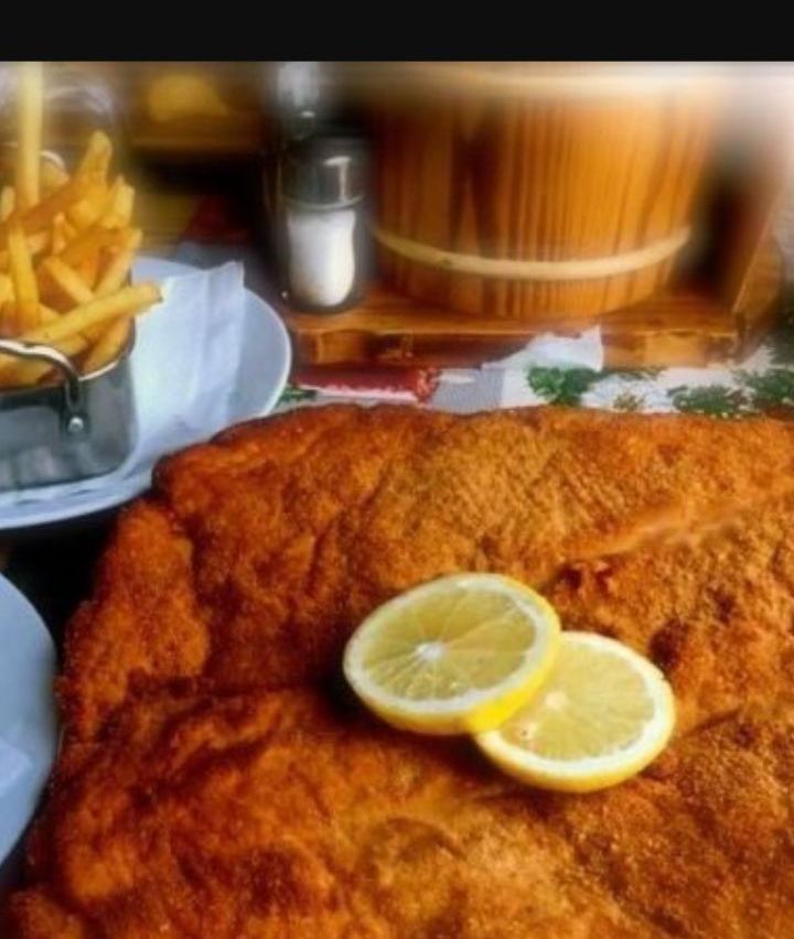 Ein gebratener Fisch mit zwei Zitronenscheiben steht auf einem Teller. Es gibt Pommes Frites in einem Behälter und ein Salzstreuer auf dem Tisch.