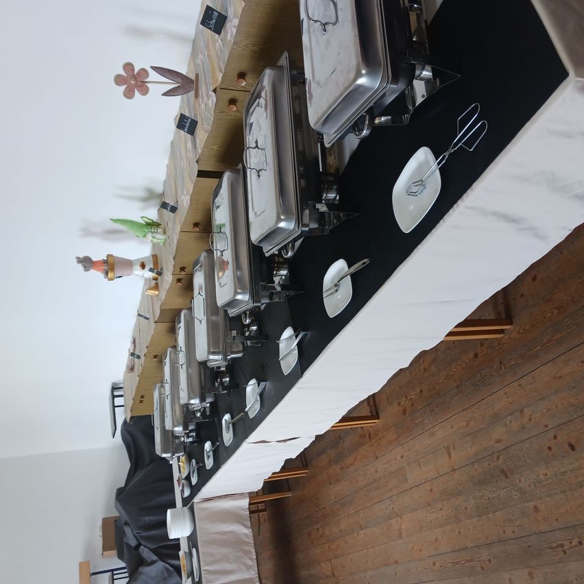 A long table covered with white cloth has several buffet containers. The containers have lids and are placed in a row. There are plates and utensils on the table. A flower and some toys are hanging from the ceiling.