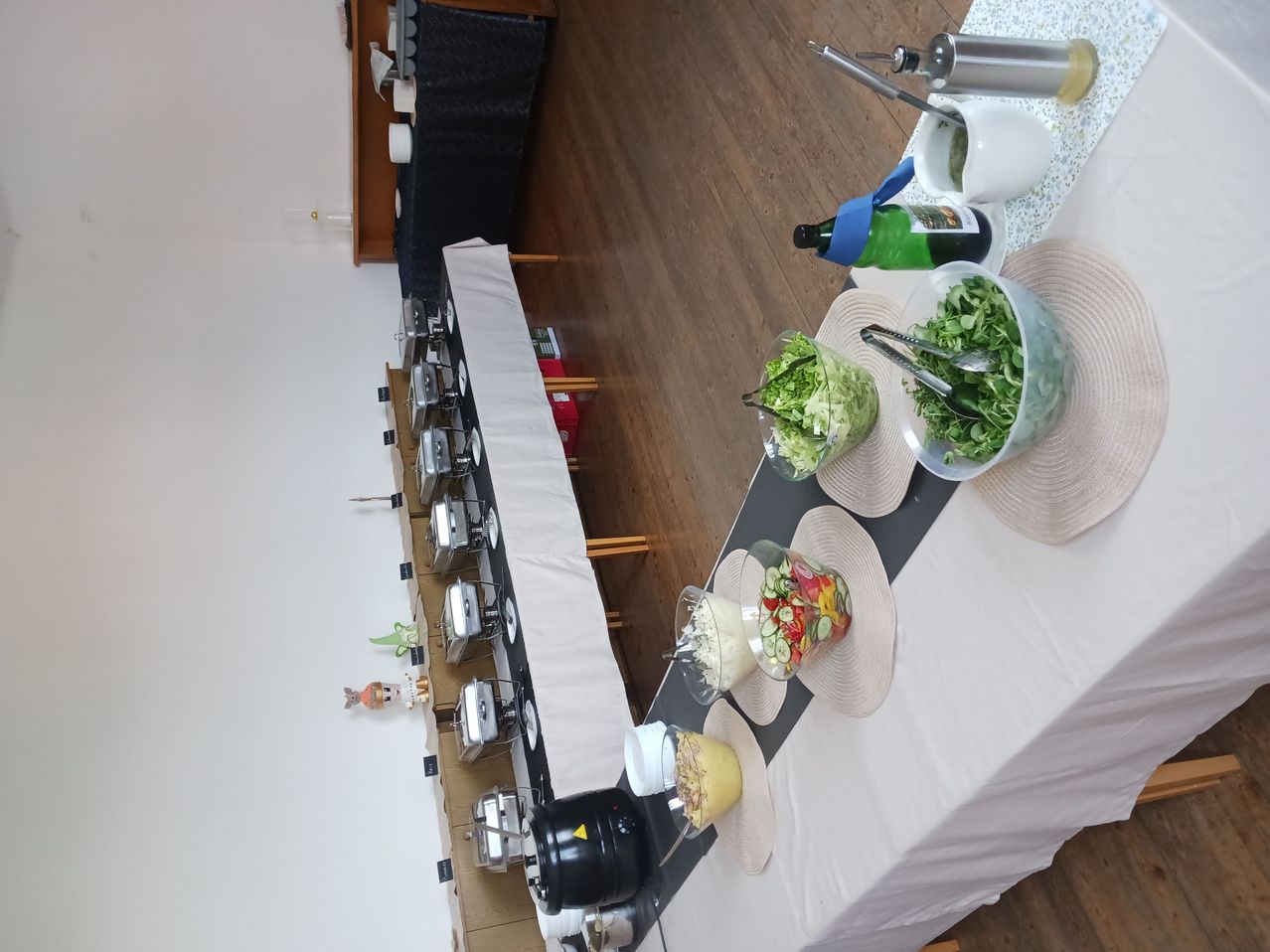 A table with a buffet setup, featuring various dishes in bowls, condiments, and salad servers. There are also food heaters and a wooden floor.