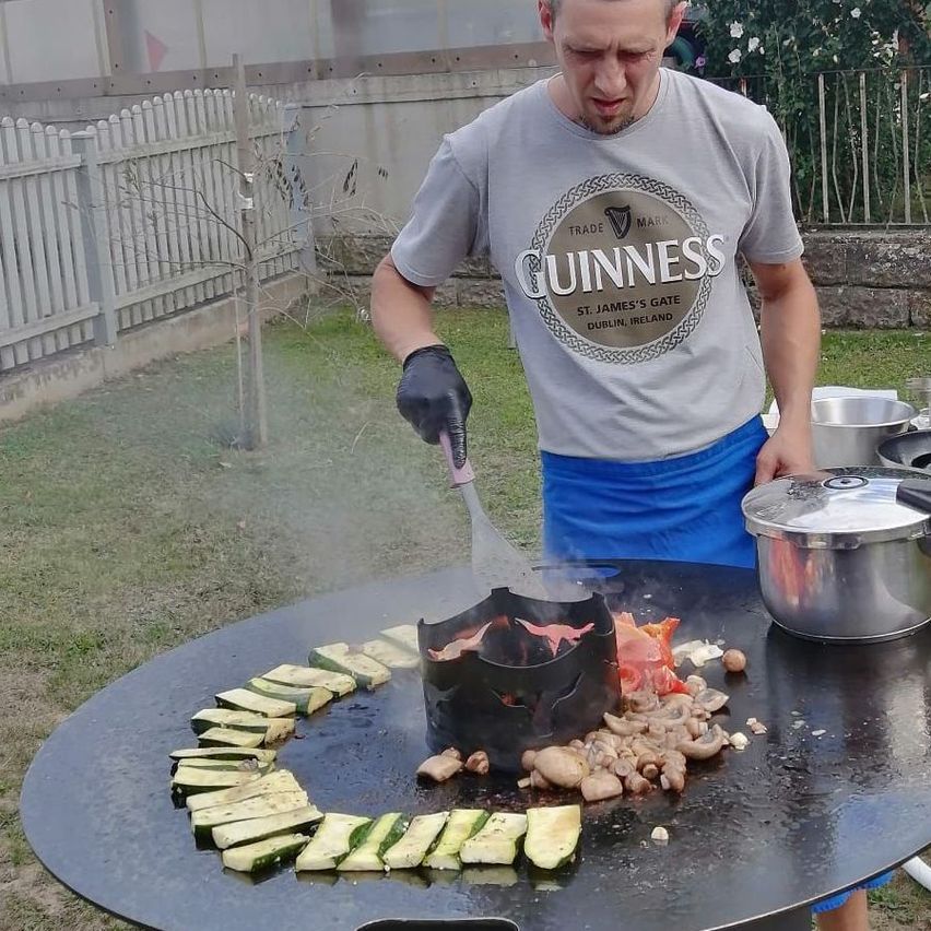 Ein Mann in einem Guinness-Shirt grillt Gemüse auf einem großen Grill im Freien.