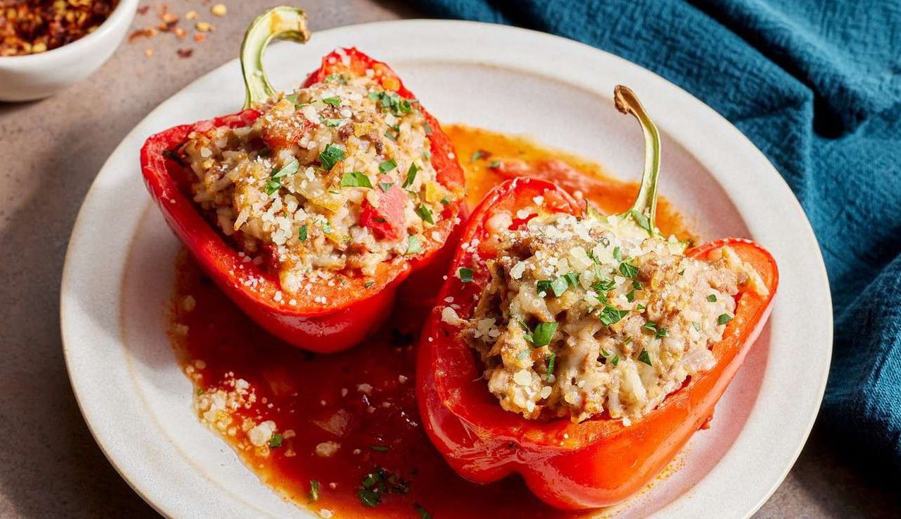 Two red bell peppers filled with meat, cheese, and rice on a white plate. The peppers are topped with herbs and sprinkled with grated cheese.