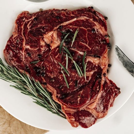 Ein roher Steak mit schwarzem Pfeffer und Rosmarin auf einem weißen Teller, begleitet von einem Messer an der Seite.