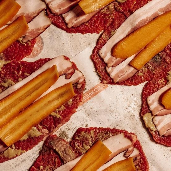 Nahaufnahme mehrerer runder Fleischpatties, belegt mit Speck und gelbem Senf, auf Pergamentpapier.