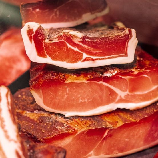 Eine Nahaufnahme von mehreren Scheiben luftgetrocknetem Fleisch, möglicherweise Prosciutto, die aufeinander gestapelt sind. Das Fleisch hat eine rötlich-rosa Farbe mit sichtbarem Fett und einer leicht knusprigen Textur.