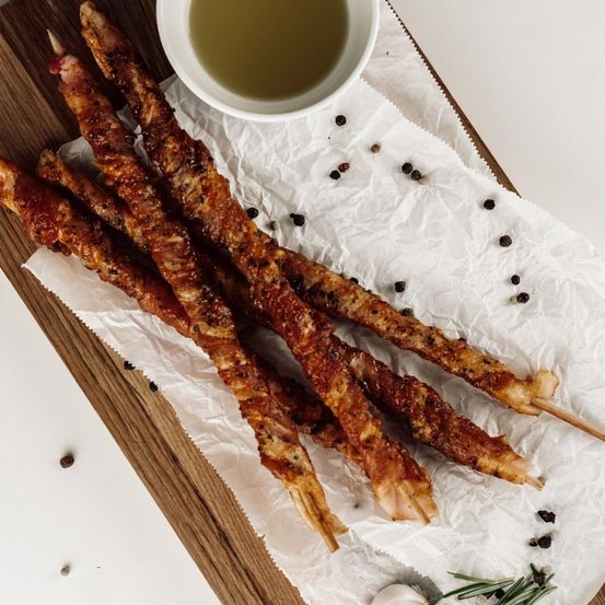 Ein Holzbrett zeigt mehrere mit Speck umwickelte Spieße, garniert mit Kräutern und Pfeffer, begleitet von einem kleinen Ölschälchen.