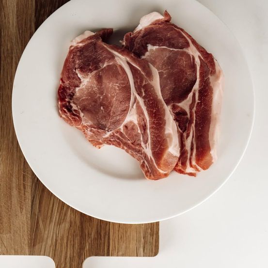 Ein roher Schweinekotelett liegt auf einem weißen Teller vor einem hölzernen Hintergrund. Das Fleisch ist roh und sieht frisch aus.