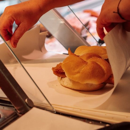 Eine Person legt ein frittiertes Hähnchensandwich auf ein weißes Papier in einer Glasvitrine.