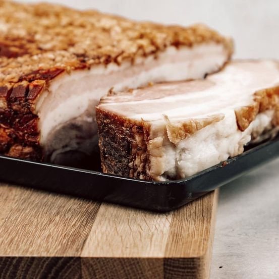 Eine Nahaufnahme eines geschnittenen Schweinebauchs mit knuspriger Kruste, auf einem Holzschneidbrett. Das Fleisch ist in dicke Stücke geschnitten und zeigt das weiße Fett und das rosa Fleisch.