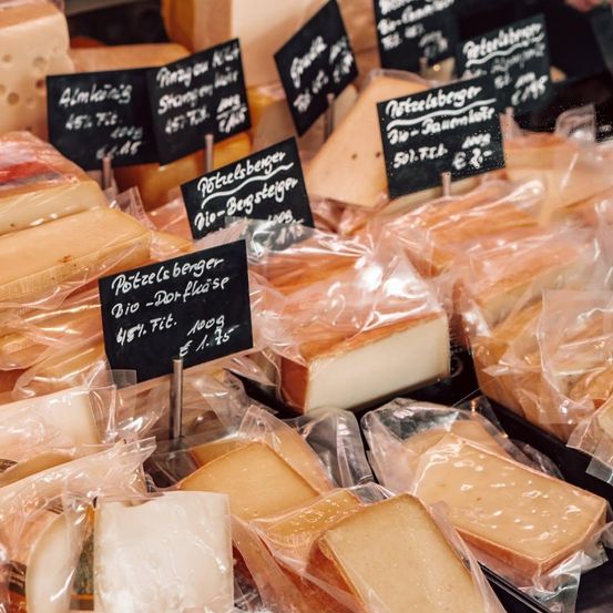 Eine Auswahl an Käsesorten in Plastikverpackungen mit Preisschildern auf einem Marktstand. Die Käsesorten sind in Reihen angeordnet, einige sind mit 'Potzels berger' und 'Bio-Dort-Fläse' beschriftet.