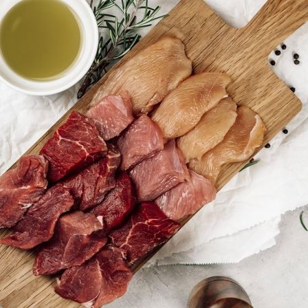 Ein Holzschneidebrett mit verschiedenen Fleischstücken, einschließlich Rind- und Hähnchenfleisch, begleitet von einer Schüssel Öl und Kräutern auf einem weißen Tuch.