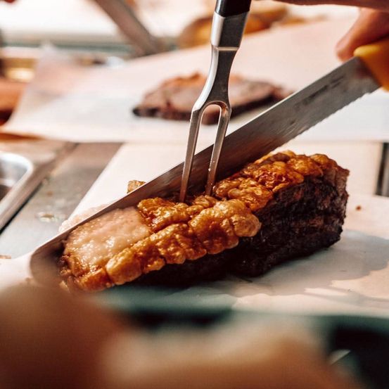 Eine Person schneidet ein Stück Fleisch mit Messer und Gabel auf einem Schneidebrett. Das Fleisch hat eine knusprige, goldbraune Kruste.