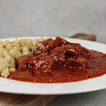Ein weißer Teller mit einer herzhaften Portion Rindergulasch, überzogen mit einer reichhaltigen roten Sauce, begleitet von cremigen Kartoffelpüree.