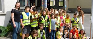 Eine Gruppe von Kindern in gelben Sicherheitswesten posiert für ein Foto vor einem kleinen Bibliotheksgebäude.