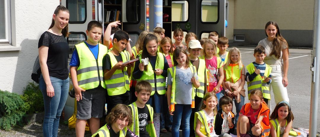 Eine Gruppe von Kindern in gelben Sicherheitswesten posiert für ein Foto vor einem kleinen Bibliotheksgebäude.