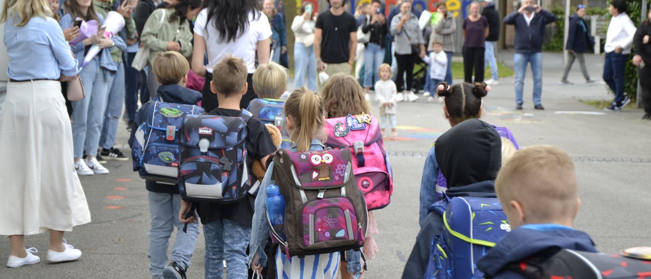 Eine Gruppe von Kindern mit Rucksäcken geht durch einen Schulhof. Hinter ihnen steht eine Menge Erwachsener und Kinder und macht Fotos. Im Hintergrund ist ein Gebäude mit farbenfroher Fassade zu sehen.