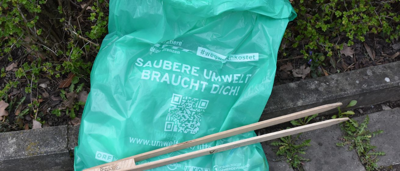 Ein grüner Plastikbeutel mit dem Text Saubere Umwelt Braucht Dich liegt neben hölzernen Chopsticks auf einer Betonfläche. Der Beutel hat einen QR-Code.