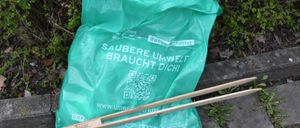 A green plastic bag with the text Saubere Umwelt Braucht Dich is beside wooden chopsticks on a concrete surface. The bag has a QR code.