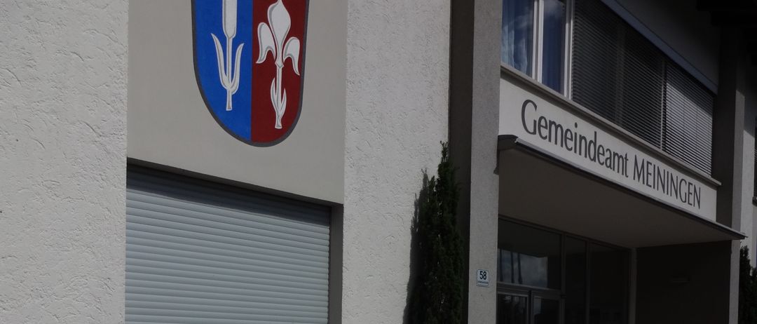 Die Vorderseite eines Gebäudes mit einem blau-weiß-roten Schild und den Worten 'Gemeindeamt Meiningen'. Darunter befindet sich ein Fenster mit einem Rollladen. Vor dem Gebäude stehen Pflanzen und Blumen.