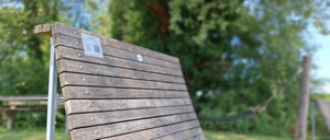 Eine Holzbank mit Metallrahmen und einem weißen Etikett mit QR-Code an der Rückenlehne. Die Bank befindet sich in einem Park mit Bäumen und Gras im Hintergrund.