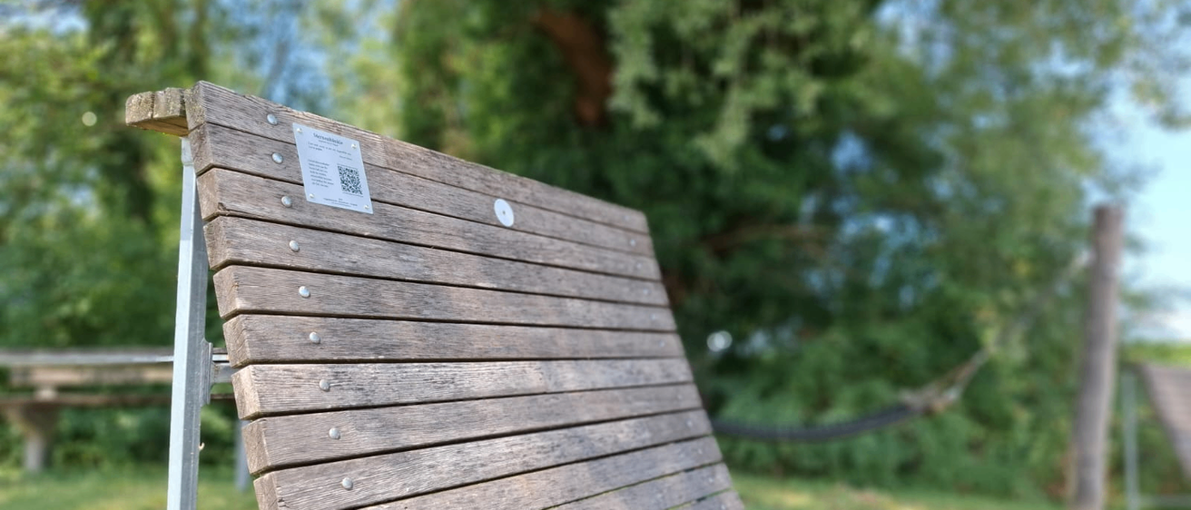 Eine Holzbank mit Metallrahmen und einem weißen Etikett mit QR-Code an der Rückenlehne. Die Bank befindet sich in einem Park mit Bäumen und Gras im Hintergrund.