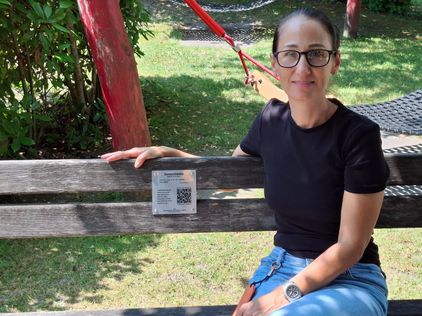 Eine Frau mit Brille und schwarzem T-Shirt sitzt auf einer Holzbank in einem Park. Ein Schild mit QR-Code ist an der Bank befestigt. Hinter ihr befindet sich eine rote Schaukel und ein grüner Rasen.