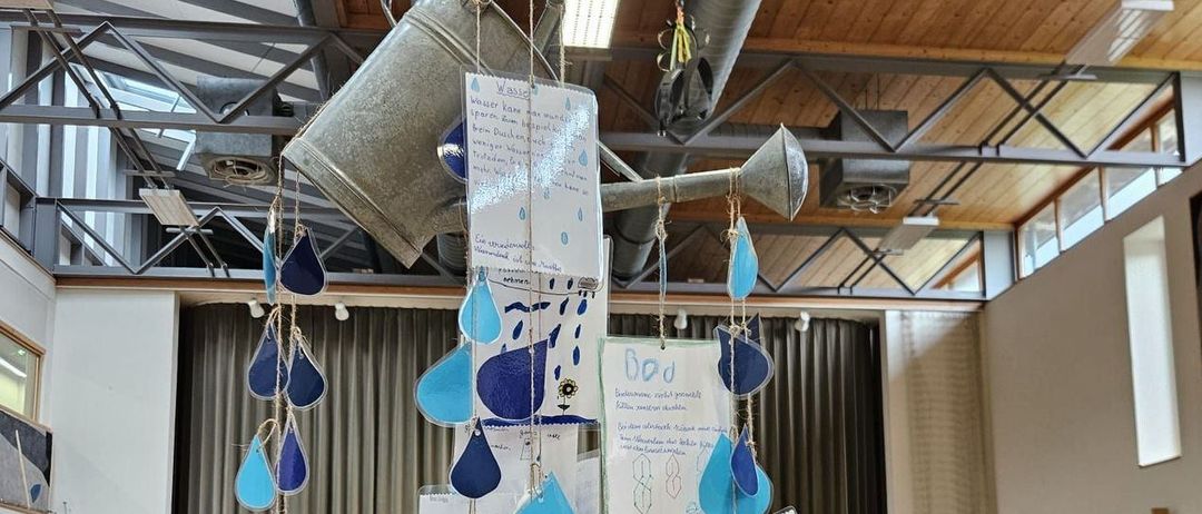 Ein Raum mit Holzstühlen, einer weißen Wand und einer Kunstinstallation mit blauen Regentropfen und einem silbernen Gießkannen, die von der Decke hängen. Es gibt Tische, Stühle und eine Tafel.