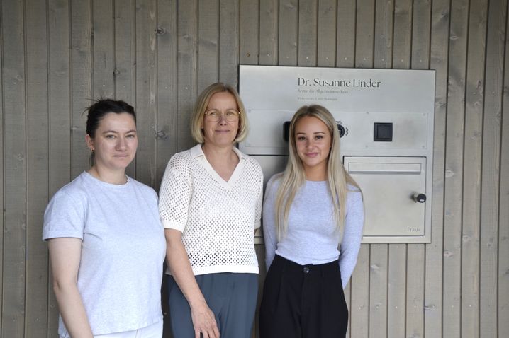 Drei Frauen stehen lächelnd vor einer Holzwand. Die Frau links trägt ein graues Shirt, die mittlere Frau eine weiße Bluse und die rechte Frau ein graues Top. Ein Schild mit der Aufschrift Dr. Susanne Linder steht hinter ihnen.