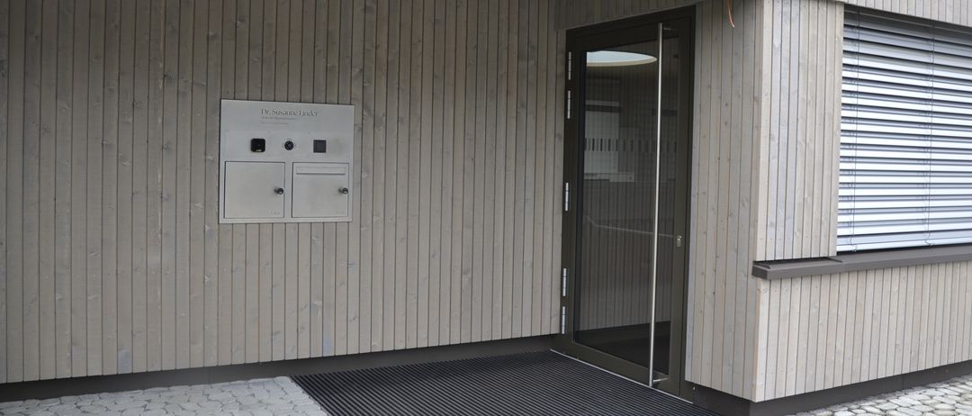 Der Eingang eines Gebäudes mit einer Glastür und einem Teppich davor, die Wand besteht aus Holz, und es gibt ein Schild mit dem Text Dr. Susanne Linda.