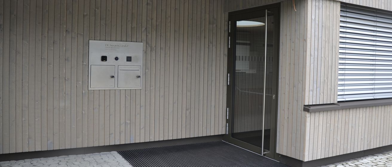 Der Eingang eines Gebäudes mit einer Glastür und einem Teppich davor, die Wand besteht aus Holz, und es gibt ein Schild mit dem Text Dr. Susanne Linda.