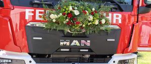 Die Front eines MAN-LKWs mit einer rot-weißen Blumenarrangement, grünen Blättern und dem Kennzeichen FW 268 FK. Das Wort 'MAN' ist prominent auf dem vorderen Stoßfänger angebracht.