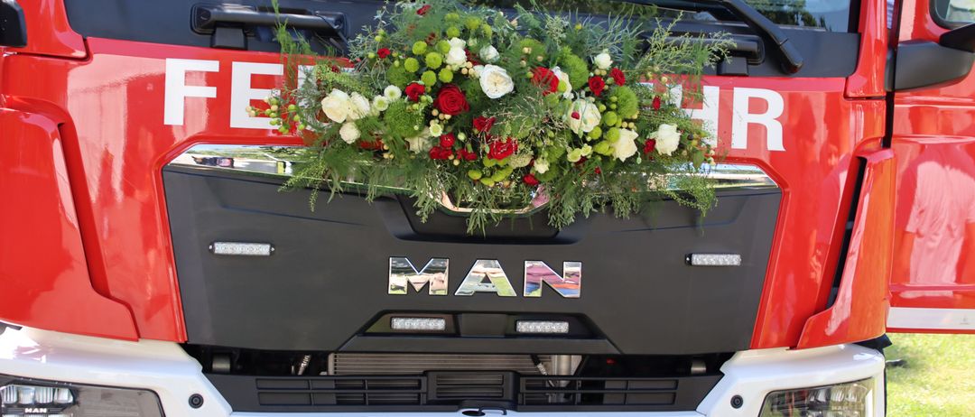 Die Front eines MAN-LKWs mit einer rot-weißen Blumenarrangement, grünen Blättern und dem Kennzeichen FW 268 FK. Das Wort 'MAN' ist prominent auf dem vorderen Stoßfänger angebracht.