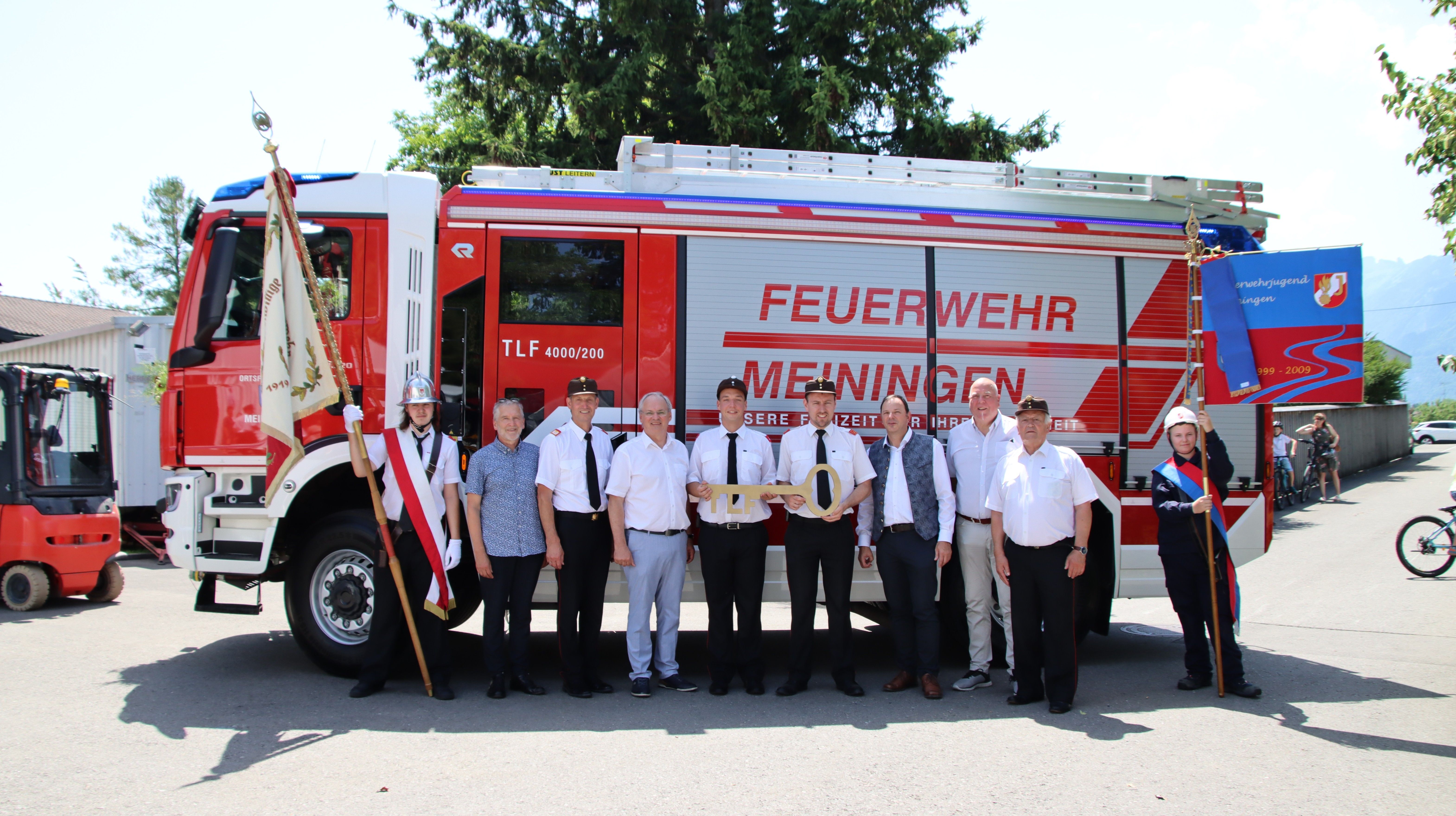 Eine Gruppe von Männern in Uniformen steht vor einem Feuerwehrauto mit der Aufschrift 'Feuerwehr Meiningen'. Sie halten einen Schlüssel in Form einer '0'. Bäume und ein heller Himmel sind im Hintergrund.