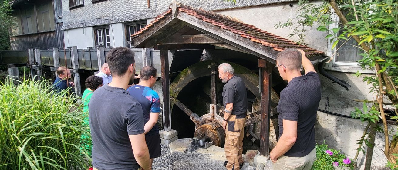 Eine Gruppe von Menschen steht um ein großes Rad in einem Schuppen herum, einer Mann lehnt sich an eine Wand. Der Schuppen ist von Pflanzen und Blumen umgeben.