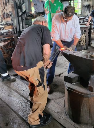 Zwei Männer in einer Werkstatt mit Metallwerkzeugen. Einer hält einen Hammer und arbeitet an einem Metall Amboss. Ein anderer Mann beobachtet. Dahinter sind Regale mit Metallwerkzeugen und Ausrüstung gefüllt.