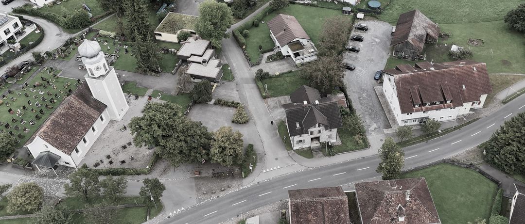 Eine Luftaufnahme eines Wohngebiets mit Häusern, Einfahrten und geparkten Autos. Die Häuser haben braune Dächer und weiße Wände, umgeben von Grünflächen. Das Gebiet ist gut gepflegt mit Bäumen und Büschen.