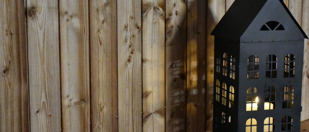 Eine dunkle, hohe Laterne steht vor einer Holzwand. Die Laterne hat viele Fenster und ist von innen beleuchtet. Die Holzwand hat Äste und Nagelspuren.