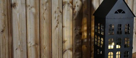 Eine dunkle, hohe Laterne steht vor einer Holzwand. Die Laterne hat viele Fenster und ist von innen beleuchtet. Die Holzwand hat Äste und Nagelspuren.