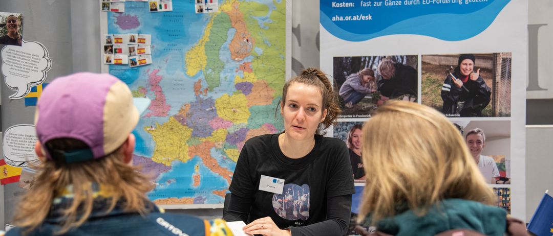 Drei junge Menschen sitzen an einem Tisch, einer spricht. Eine Karte von Europa ist im Hintergrund. Die Frau in der Mitte trägt ein schwarzes T-Shirt.