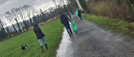 Mehrere Menschen gehen mit grünen Tüten in der Hand auf einem nassen Pfad in einem Grasfeld. Sie scheinen an einer Müllsammelaktion teilzunehmen. Ein schwarz-weißer Hund folgt ihnen. Das Gebiet ist von Bäumen umgeben.