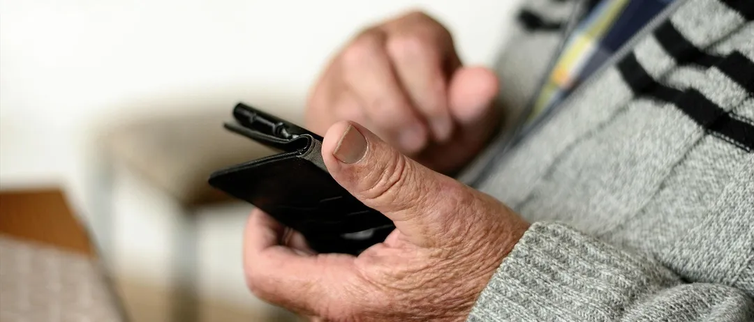 Ein Nahaufnahme der Hände einer älteren Person, die ein schwarzes Smartphone hält, trägt einen grauen Pullover.